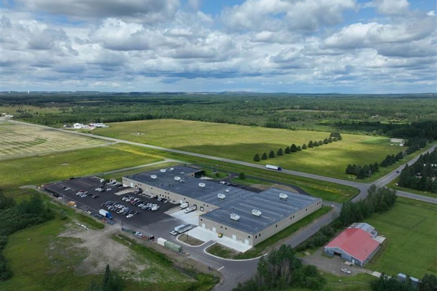 Aerial view of large industrial building and parking lot surrounded by green fields under a cloudy sky.