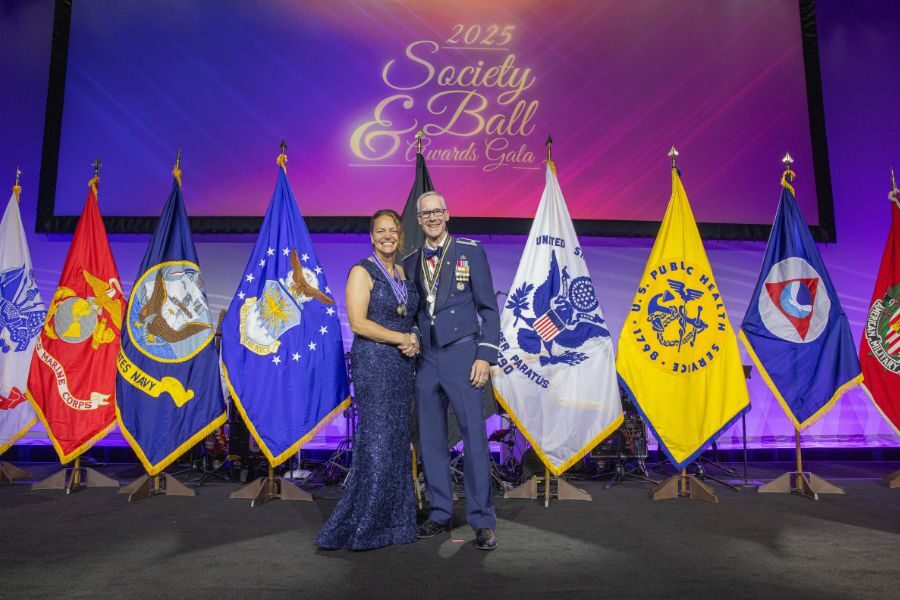Man and woman smiling, wearing medals, in front of military flags. 