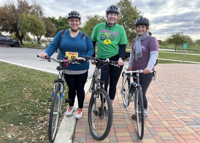 Three cyclists stand smiling with bikes, wearing helmets and casual athletic clothing, in a park on a cloudy day.
