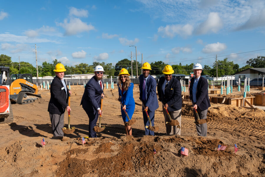 Dawn Aiello and five colleagues, wearing helmets, participate in a groundbreaking ceremony, shoveling dirt at a construction site.