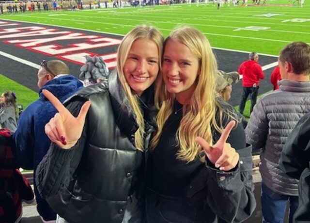 Two women smiling, posing with fingers in a "guns up" gesture at a sports stadium.