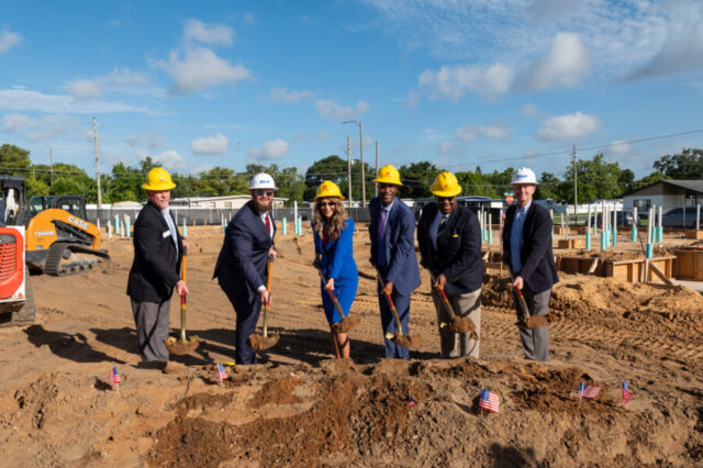 Dawn Aiello and five colleagues, wearing helmets, participate in a groundbreaking ceremony, shoveling dirt at a construction site.